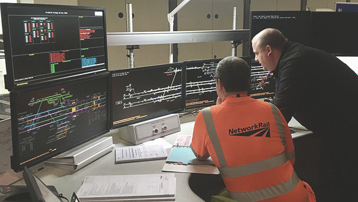 Inside Manchester Rail Operations Centre. Two men in front of many computer screens. One is wearing a Network Rail orange hi-viz jacket.