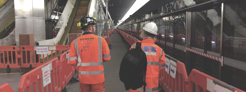 Elizabeth line during construction with 2 people walking between barriers on an underground station platform. One is wearing a ricardo logo on their hi-viz jacket.
