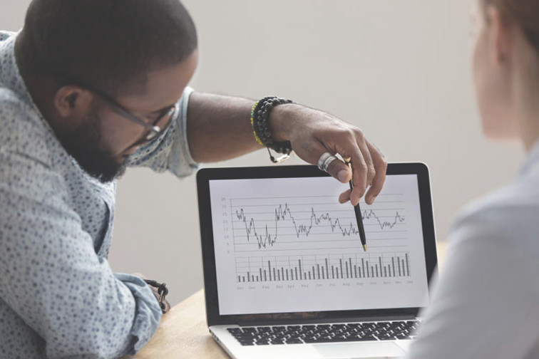 Man pointing at a graph on a laptop screen with a pen whilst another person looks from the foreground