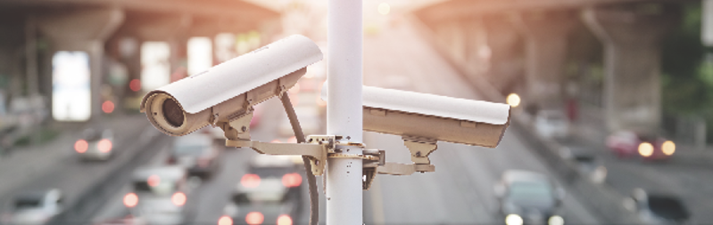 Pole mounted CCTV cameras above a large city road with cars on it in the background.